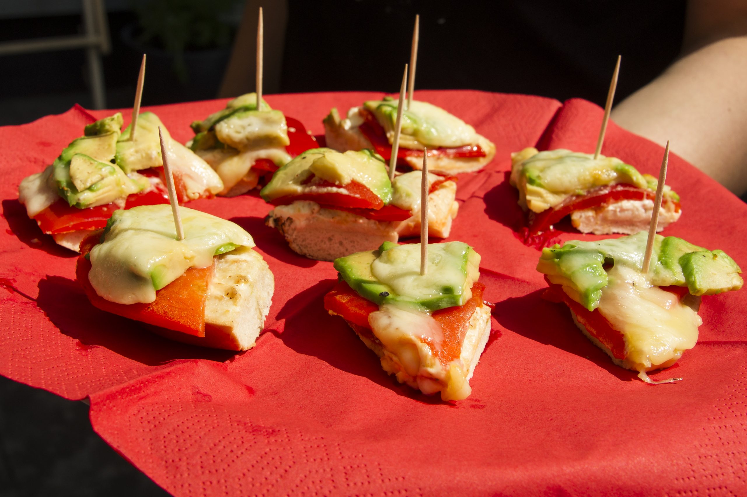 Genießen Sie die Erfahrung der Erlebnisgastronomie mit mundgerechten Häppchen: Käse, Avocado und rote Paprika, elegant auf Brot angerichtet und mit Zahnstochern auf einer schicken roten Serviette aufgespießt.