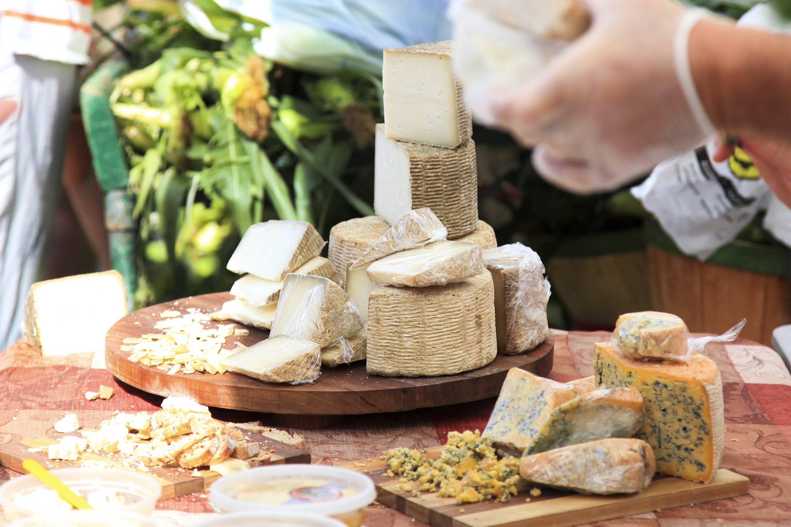 Verschiedene Käsesorten auf einer Holzplatte, während Hände auf einem Markt im Freien Proben vorbereiten und so die Essenz der Erlebnisgastronomie einfangen.