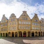 Historischer europäischer Platz mit mittelalterlichen Gebäuden, Kopfsteinpflaster und einem hohen Kirchturm unter einem blauen Himmel.