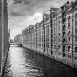 Ein Schwarzweißfoto zeigt die historische Speicherstadt in Hamburg mit einem Kanal, gesäumt von alten Backsteingebäuden unter einem bewölkten Himmel.