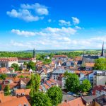 Malerische Aussicht auf eine Stadt mit Gebäuden mit roten Dächern, einer Kirche und Bäumen unter einem blauen Himmel mit Wolken.