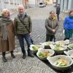 Fünf Menschen stehen auf einer Kopfsteinpflasterstraße und bewundern ein Tablett mit Tellergerichten, jedes davon so hübsch angerichtet, als handele es sich um ein wertvolles Mitarbeitergeschenk.