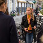 Eine Frau mit einem Schlüsselband spricht auf einer Stadtstraße in der Nähe geparkter Fahrräder zu einer Gruppe von Menschen.