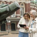 Zwei lächelnde Frauen bedienen ein Tablet und nehmen an einem Stadtquiz teil, während sie vor den beeindruckenden Skulpturen und der historischen Architektur Wiesbadens im berühmten Historischen Fünfeck stehen.