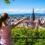 Eine Frau zeigt auf eine Stadtansicht mit einem markanten Kirchturm, umgeben von üppigem Grün unter einem klaren Himmel, was sie an eine Illustration aus dem letzten Newsletter erinnert.