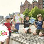 Vier Kinder sitzen auf einer Bank in einer historischen Stadtkulisse und lesen ihre GEOlino-Zeitschriften. Inmitten der lebendigen Kulisse der Hamburger HafenCity genießen sie ihr Outdoor-Abenteuer Stadtrallye.