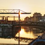 Sonnenuntergang über einem Industriehafen mit Kränen, Containern und einer Brücke, die sich im ruhigen Wasser spiegelt.