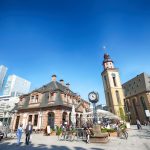 Stadtplatz mit Menschen, historischen Gebäuden, einem Uhrenturm und modernen Wolkenkratzern im Hintergrund an einem sonnigen Tag.