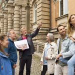 Die Reisegruppe hört ihrem Reiseleiter aufmerksam zu, während sie während einer fesselnden Historischen Tour in Berlin auf den Stufen des Friedrichswerders steht und sitzt, einem Ort, der für seine historische Fassade bekannt ist.