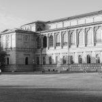 Schwarzweißfoto eines großen historischen Gebäudes im Herzen der Maxvorstadt, München, mit Menschen, die auf einer Wiese davor entspannen. Perfekt für alle, die an einer historischen Tour teilnehmen möchten, um die reiche Geschichte und den Charme dieses berühmten Viertels zu entdecken.