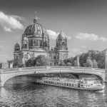 Schwarz-Weiß-Bild eines Flusskreuzfahrtschiffs, das unter einer Brücke in der Nähe des großen Kuppeldoms in Berlin durchfährt und an den historischen Charme von Friedrichswerder erinnert.