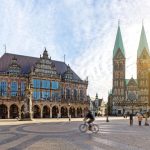 Ein Radfahrer fährt unter einem sonnigen Himmel über einen historischen europäischen Platz mit reich verzierten Gebäuden und zwei Kirchtürmen.