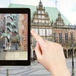 Hände halten eine Tafel mit der Statue der Bremer Stadtmusikanten, im Hintergrund ist ein historisches Gebäude zu sehen.