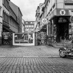 Eine Straßenszene im Herzen von St. Pauli mit Kopfsteinpflaster, das zu einem geparkten Motorrad führt, und einer mit Graffiti geschmückten Gasse fängt die Essenz einer historischen Tour durch Hamburgs lebendige Vergangenheit ein – alles in eindrucksvollem Schwarzweiß.