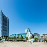 Panoramablick auf moderne Gebäude, darunter ein hoher Turm und Glaskonstruktionen, unter einem klaren blauen Himmel.