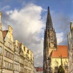 Eine historische Kirche mit einem hohen Turm steht zwischen alten Gebäuden unter einem wolkig-blauen Himmel.