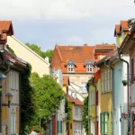 Eine schmale Straße mit farbenfrohen Häusern, schrägen Dächern und viel Grün genießt die Wärme eines sonnigen Tages.