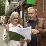 Ein älteres Ehepaar hält während seiner Rätseltour im Freien neben einem Backsteingebäude in einer belebten Stadtstraße inne und überprüft sorgfältig Dokumente.