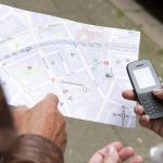 Zwei Menschen stehen, vertieft in ihre Rätseltour, mit einer Karte und einem Telefon in der Hand nachdenklich auf dem Bürgersteig.