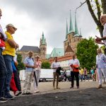 Eine Gruppe von Menschen hört einem Reiseführer in der Nähe eines historischen Gebäudes mit Türmen unter einem bewölkten Himmel zu.