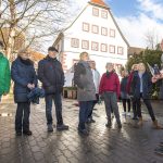 Eine Gruppe von Menschen steht an einem sonnigen Tag draußen vor einem traditionellen Gebäude und hört einem Reiseführer zu.