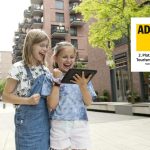 Zwei aufgeregte Mädchen mit einem Tablet in einer urbanen Umgebung neben einem Banner des ADAC Tourismuspreises, als würden sie das ultimative Kindergeburtstagsabenteuer planen.