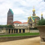 Ein Park mit einem hohen Backsteinturm und einem reich verzierten Gebäude mit goldenen Kuppeln unter einem bewölkten Himmel.