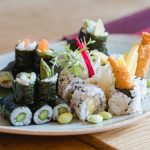 Teller mit verschiedenen Sushi-Rollen, garniert mit Gemüse, Edamame und Tempura-Garnelen auf einem Holztisch.