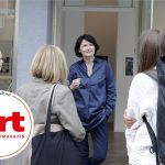 Eine Frau in blauem Outfit spricht vor einem Gebäude mit drei Personen. In der Ecke ist das Logo einer Zeitschrift zu sehen.