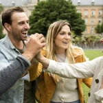 Vier Menschen lächeln und geben sich High Fives in einem Park mit Bäumen und einem großen Gebäude im Hintergrund.