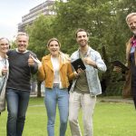 Fünf Menschen lächeln und strecken in einem Park den Daumen hoch, sie halten ein Buch und eine Tasche in den Händen.