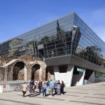 Modernes Glasgebäude mit historischen Steinruinen davor, Menschen gehen auf einem gepflasterten Weg unter einem klaren blauen Himmel.