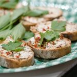 Mit cremigem Aufstrich, Kräutern und roter Paprika belegte Brotscheiben auf einem grün karierten Teller.