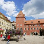 Historischer europäischer Platz mit farbenfrohen Gebäuden, einem Brunnen und Menschen, die unter einem teilweise bewölkten Himmel spazieren gehen.