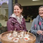 Zwei lächelnde Frauen stehen vor einem Geschäft an einem Tisch mit kleinen Schüsseln voller Desserts.