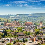 Luftaufnahme einer europäischen Stadt mit historischen Gebäuden, grünen Hügeln und blauem Himmel.