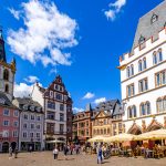 Historischer europäischer Stadtplatz mit farbenfrohen alten Gebäuden, Uhrenturm und Menschen, die unter einem blauen Himmel spazieren gehen und essen.