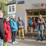 Eine Gruppe von Menschen hört einer Frau zu, die vor einem Laden mit einem „Pauli Kocht“-Schild spricht.