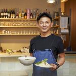 Eine Person in einer blauen Schürze hält in einem Restaurant eine Schüssel Suppe und einen Teller mit Essen.