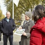 Ein Reiseleiter zeigt einer Gruppe von Menschen im Freien auf einem von Bäumen gesäumten Weg ein Foto.