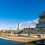 Moderne Gebäude am Flussufer unter einem klaren blauen Himmel, mit einer geschwungenen Uferpromenade und einer Brücke in der Ferne.