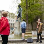 Eine Gruppe von Menschen hört einem Reiseführer in der Nähe eines Brunnens mit einer Statue in einer Parkanlage zu.