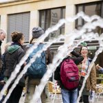 Menschen versammelten sich vor einem Gebäude. Im Vordergrund stand ein Springbrunnen, der Wasserbögen erzeugte.