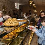 Frau serviert einem Kind und einem Erwachsenen Gebäck in einer Bäckerei mit dekorativer Beleuchtung und einer Glasvitrine.