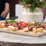 Käse- und Salami-Spieße mit Kirschtomaten auf einem Holzbrett, präsentiert auf einem Tisch im Freien.