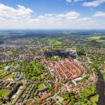 Luftaufnahme einer Stadt mit Gebäuden mit roten Dächern, grünen Feldern und einem gewundenen Fluss unter einem teilweise bewölkten Himmel.