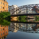 Eine Stahlbrücke über einem ruhigen Fluss spiegelt die umliegenden Backstein- und modernen Gebäude unter einem klaren Himmel wider.