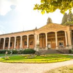 Historisches Gebäude mit Säulen und Garten davor, unter einem hellen Himmel mit Bäumen im Hintergrund.