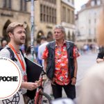 Menschen diskutieren auf einem Stadtplatz, eine Person hält einen Ordner.