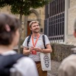 Ein Reiseführer in der Altstadt spricht vor einem historischen Gebäude zu einer Gruppe. Er hält ein rotes Schlüsselband und trägt eine Umhängetasche. Diese Historische Tour bietet Einblicke in Münsters reiche Vergangenheit und verbindet Architektur mit faszinierenden Geschichten dieser charmanten Stadt.